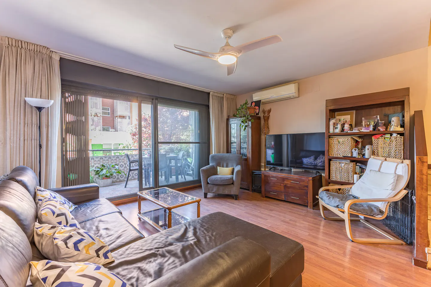 Living room with brown leather sofa, glass table, gray armchair, and wood floors. Balcony visible through sliding glass doors.
