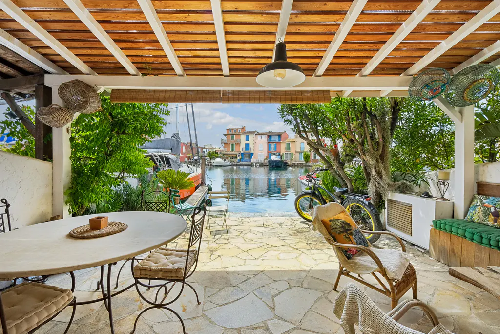 Covered patio with stone floor, table, chairs, and view of canal with colorful buildings and boats. A bicycle is parked nearby.