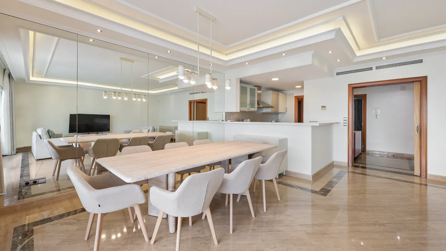 Modern dining room with a light wood table, six beige chairs, marble floors, and a mirrored wall reflecting the living area.