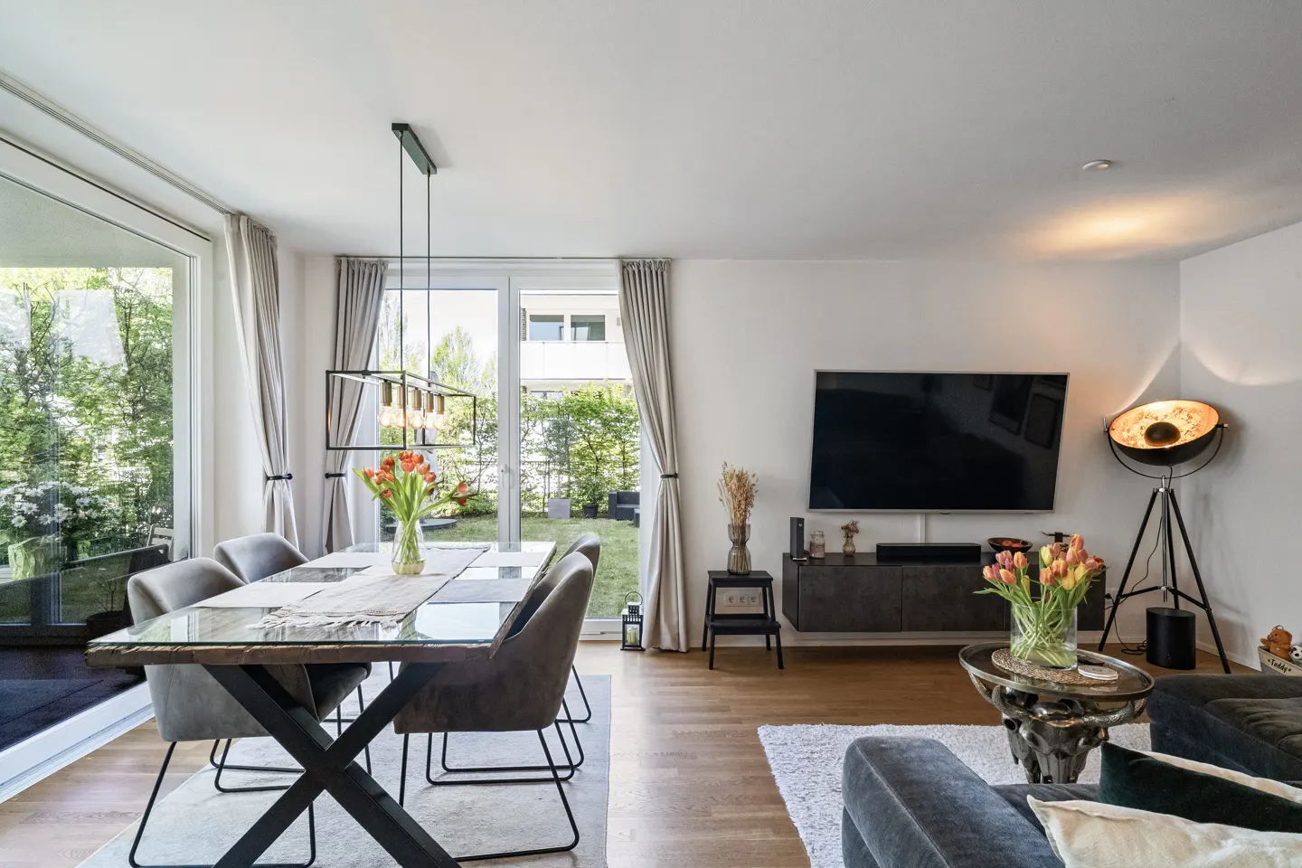 Bright, modern living and dining area with a glass-topped table, gray chairs, and a large TV. A garden is visible through the window.