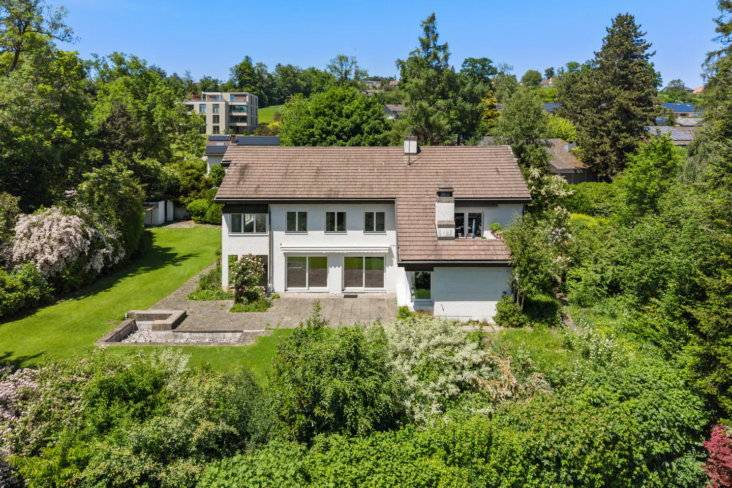 Two-story white house with a brown tiled roof, surrounded by a lush green lawn, trees, and a stone patio.