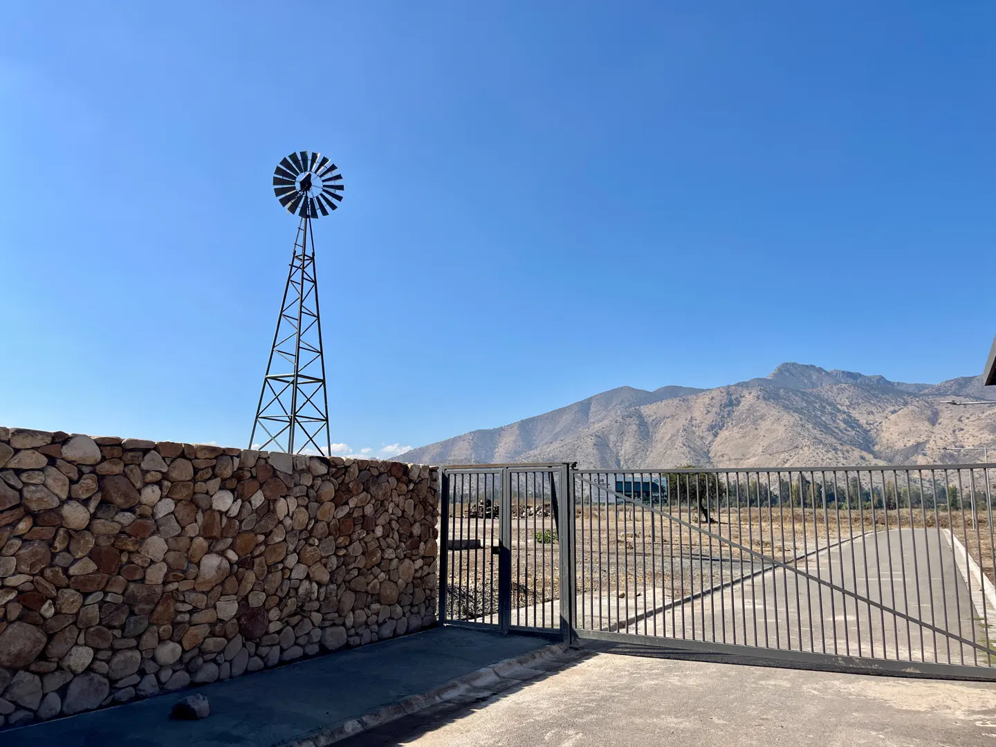 Gated property with a windmill, stone wall, and mountain backdrop under a clear blue sky.