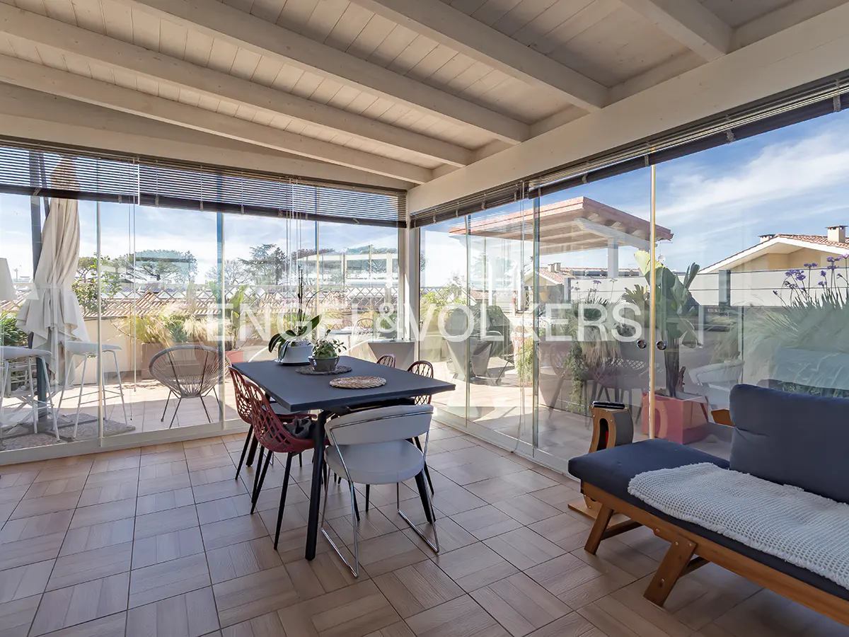 Sunroom with a dining table, chairs, and a lounge sofa, featuring glass walls overlooking a terrace with outdoor furniture.