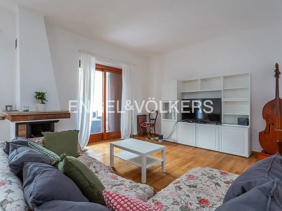 Living room with floral sofa, white coffee table, TV, and cello. Fireplace and balcony in background.