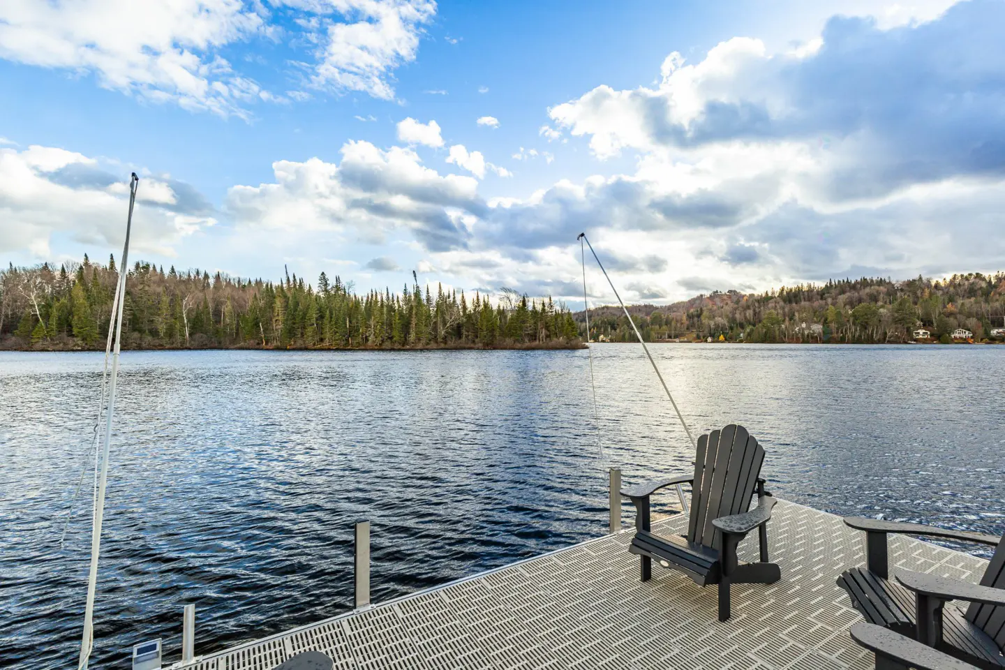 Lake view from a gray dock with two Adirondack chairs. Forest and cloudy sky in the background.