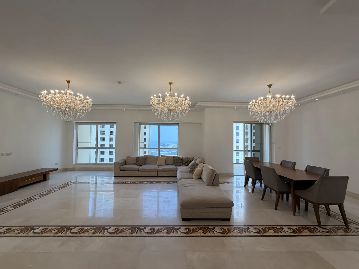 Bright, open living space with marble floors, beige sectional sofa, dining table, and three crystal chandeliers. Windows offer a city view.
