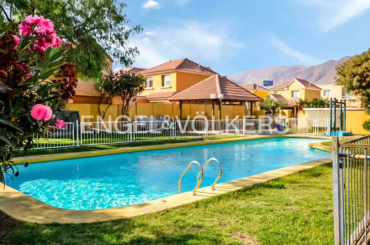 A turquoise pool is surrounded by green grass and yellow buildings under a blue sky. Engel & Völkers logo is visible.