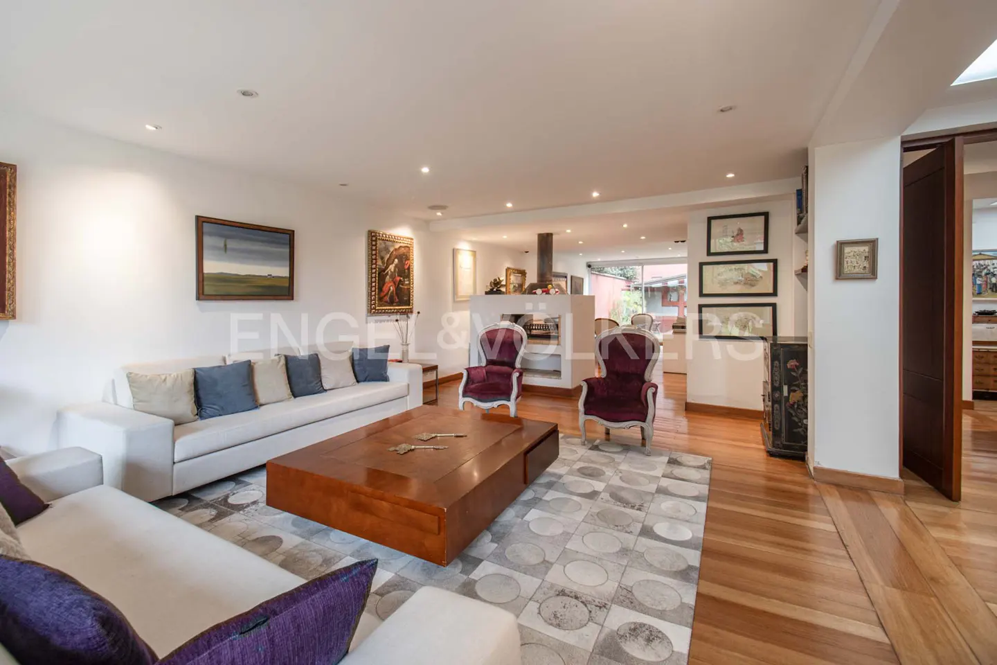 A bright living room with white sofas, a wood coffee table, and two burgundy chairs on a patterned rug. Art adorns the walls.
