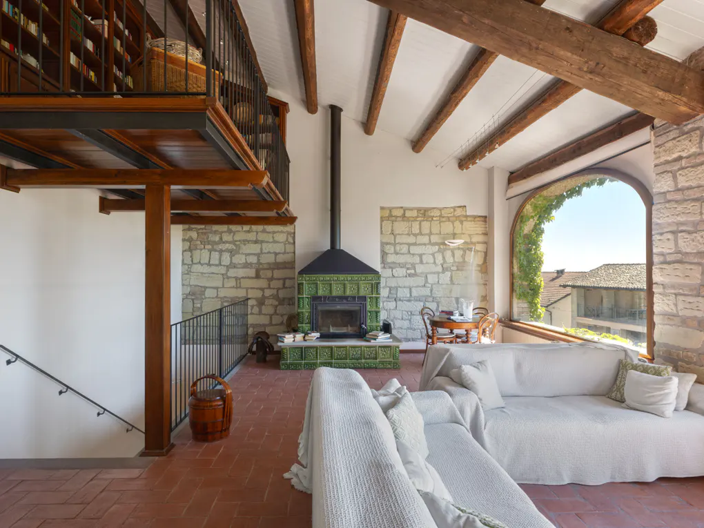 A living room with a stone wall, green tiled fireplace, white sofa, and a large arched window. A loft with a bookshelf is visible.