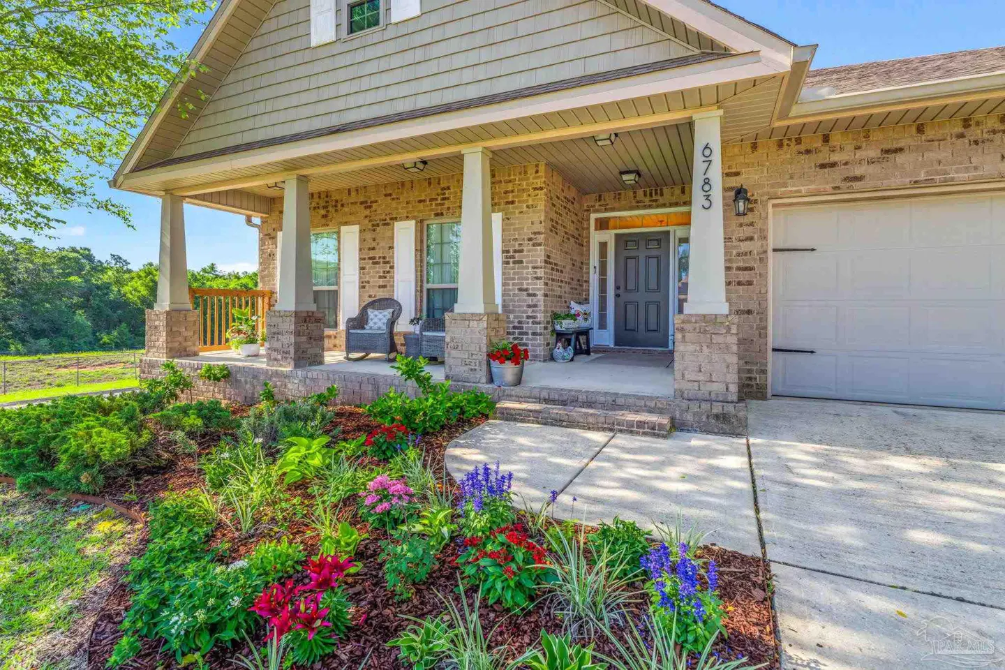 Brick house with a front porch, white pillars, and a gray front door. A flower bed with colorful flowers is in the foreground.