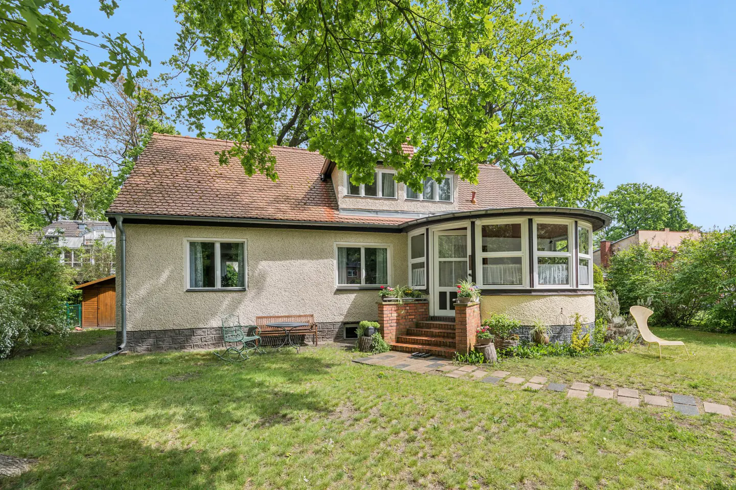 Exterior view of a beige house with a brown roof, a sunroom, and a green lawn with trees.