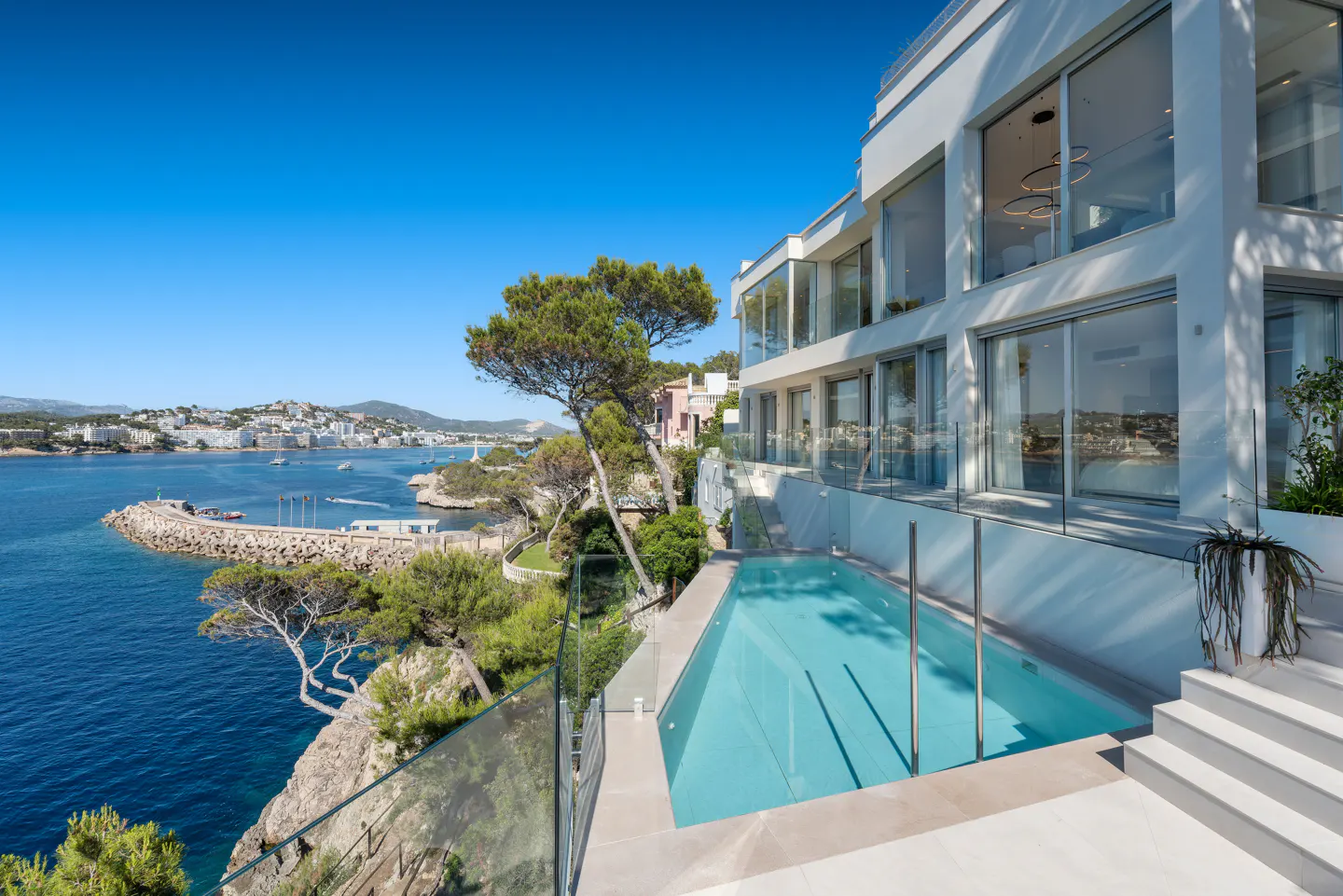 Modern white house with a pool overlooking the ocean. Clear blue sky. Trees and greenery surround the property.