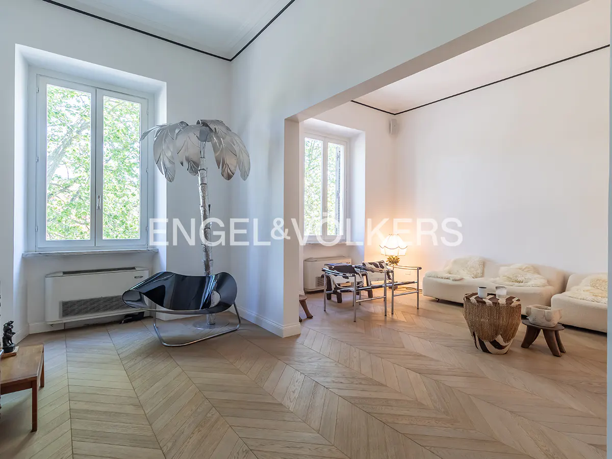 Bright living room with herringbone wood floors, white walls, and large windows. A silver palm tree lamp and black chair are near the window.