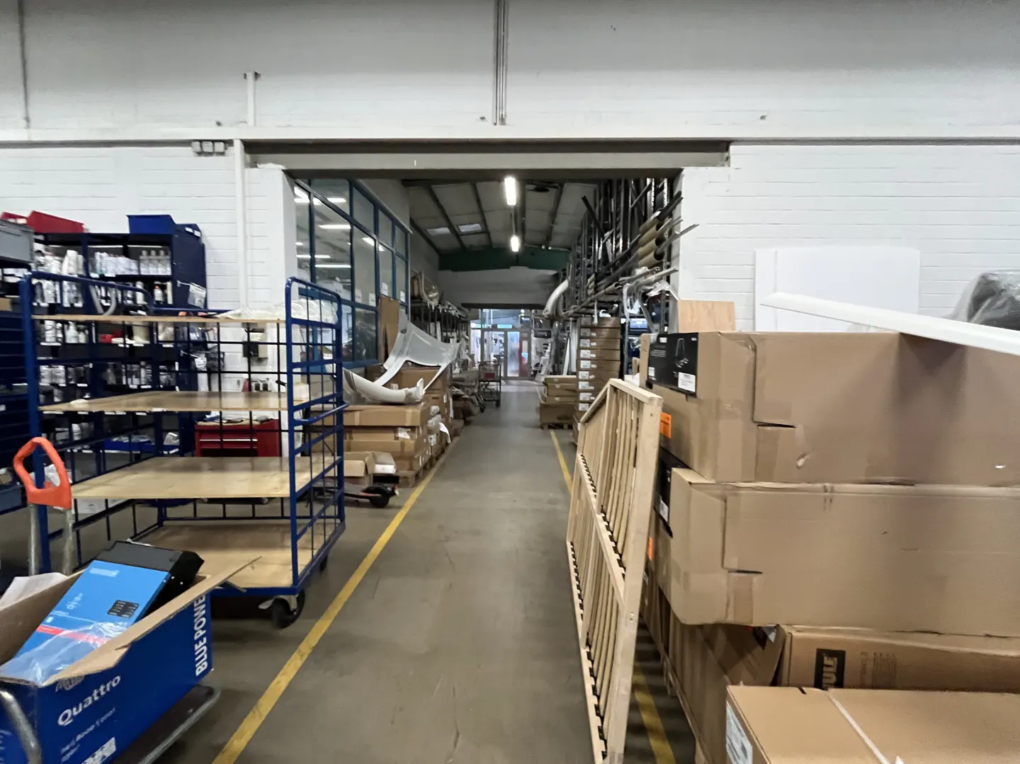A warehouse aisle with a blue metal cart on the left and stacks of cardboard boxes on the right, leading to a back exit.