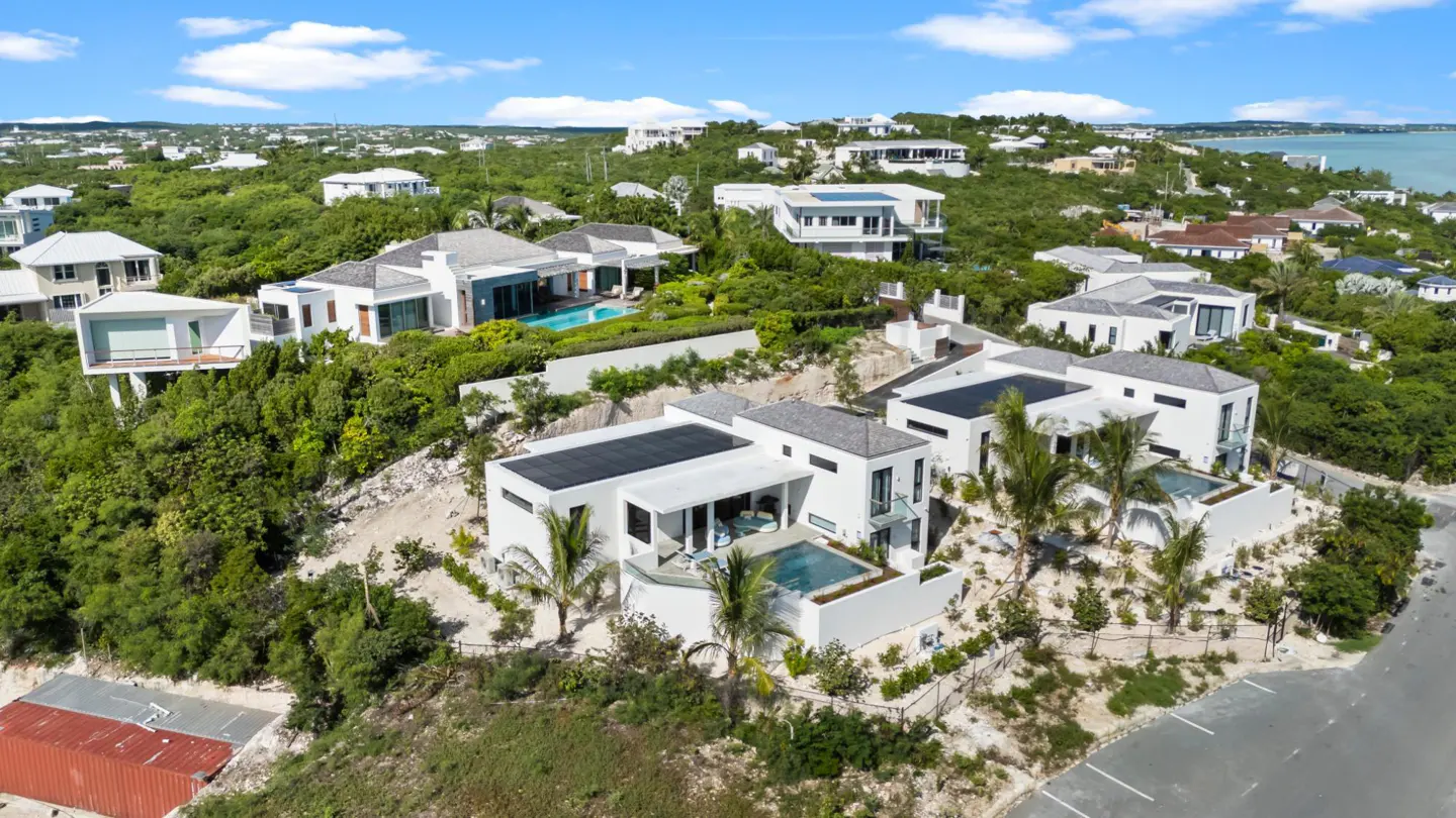 Aerial view of modern white villas with pools nestled among lush green trees, under a bright blue sky with scattered clouds.