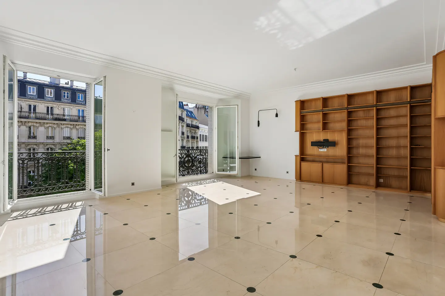 Bright, empty room with white walls, marble floors, and open French doors to balconies overlooking a Parisian street. A large wooden bookcase is on the right.