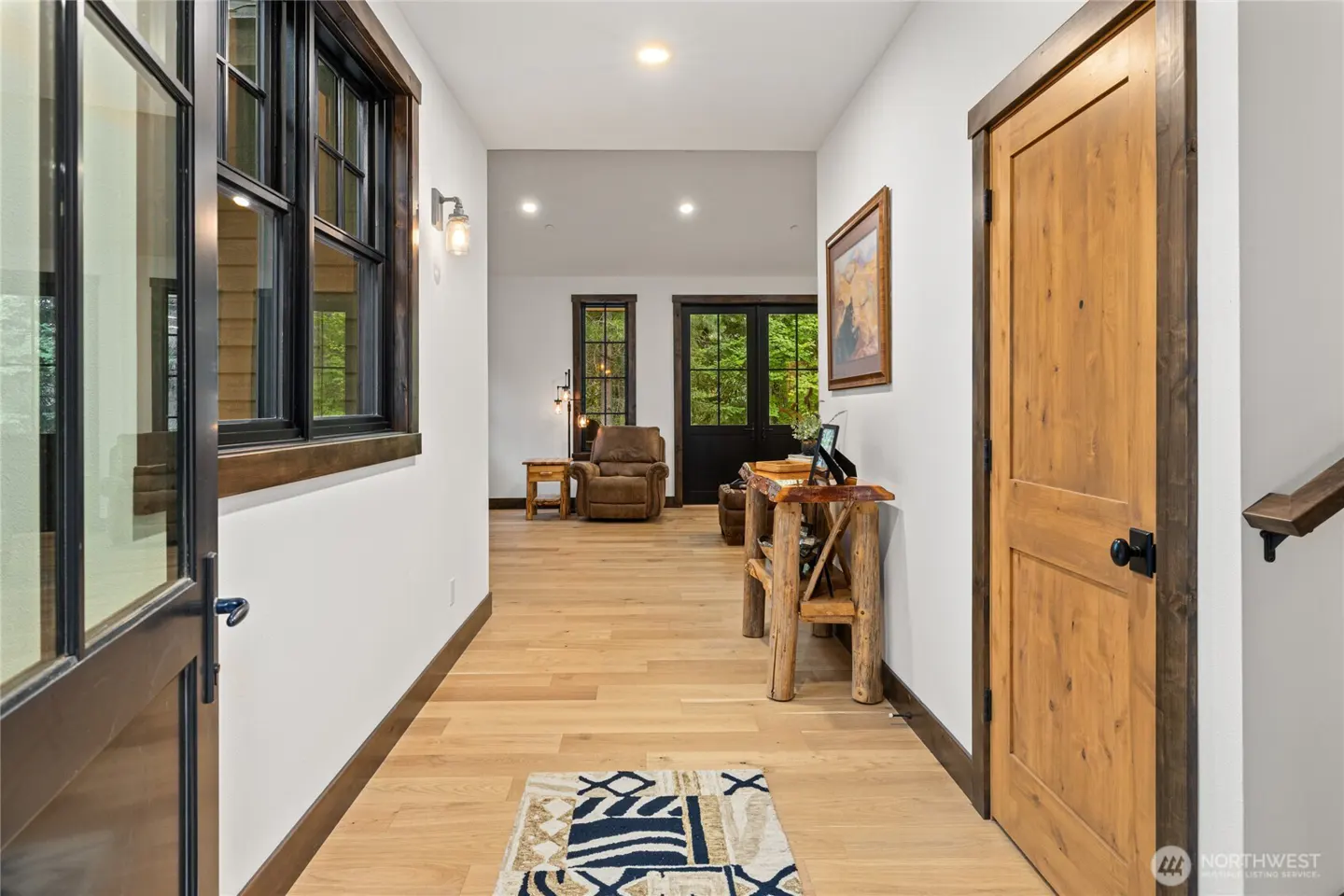 Hallway with light wood floors, white walls, and dark trim. A rustic wood table and rug accent the space. Windows and doors lead to the outside.