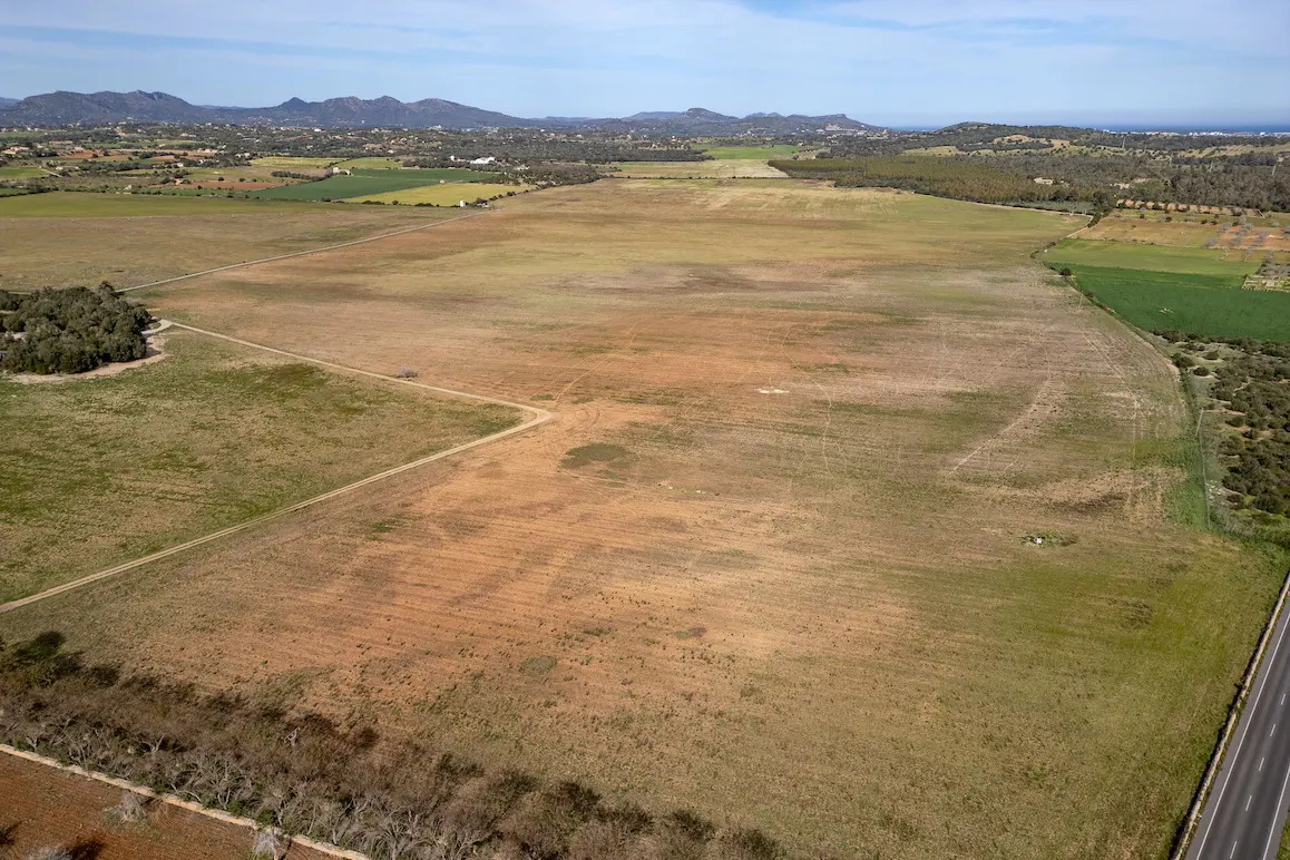Aerial view of a large, mostly brown field with some green patches, bordered by trees and a road, with mountains in the distance.