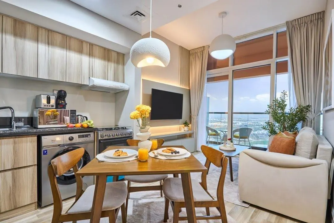 Bright apartment interior with a kitchen, dining table set for breakfast, and a living area with a balcony overlooking a cityscape.