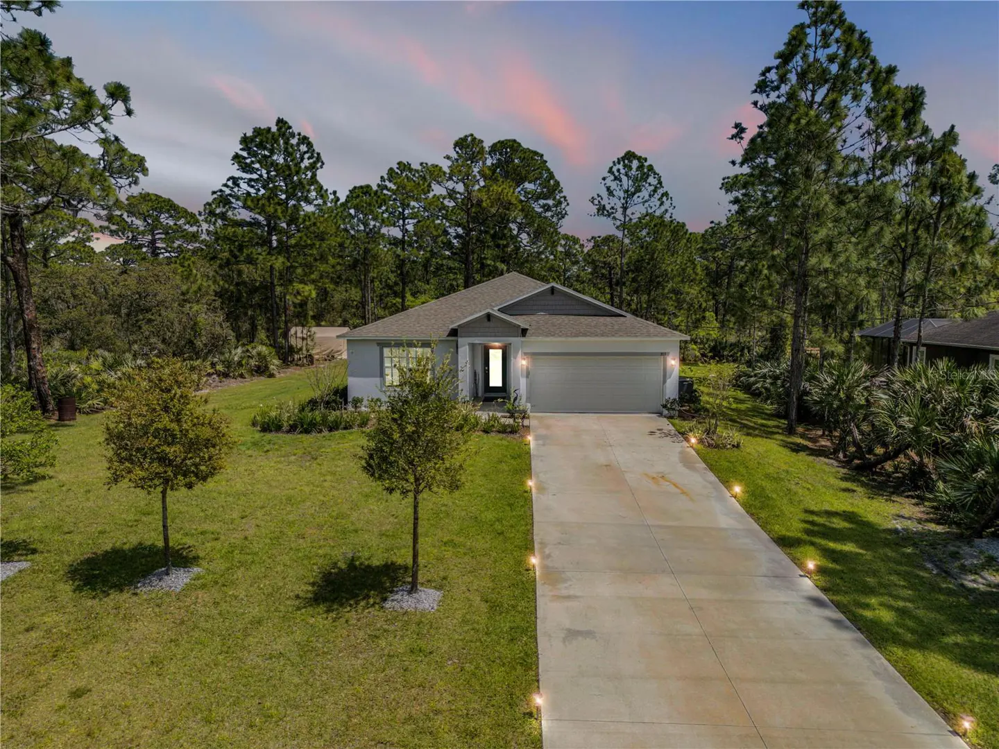 Single-story light gray home with a concrete driveway and landscape lighting at dusk, surrounded by trees and green lawn.