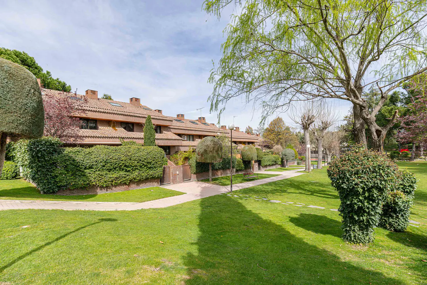 Exterior view of townhouses with red tile roofs, green lawns, hedges, trees, and a sidewalk on a sunny day.