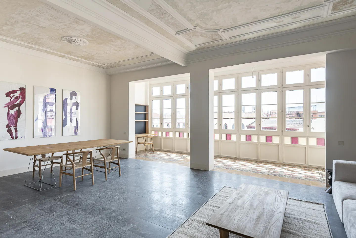 Bright, airy room with large windows, wood table and chairs, and abstract art. Ornate ceiling detail.