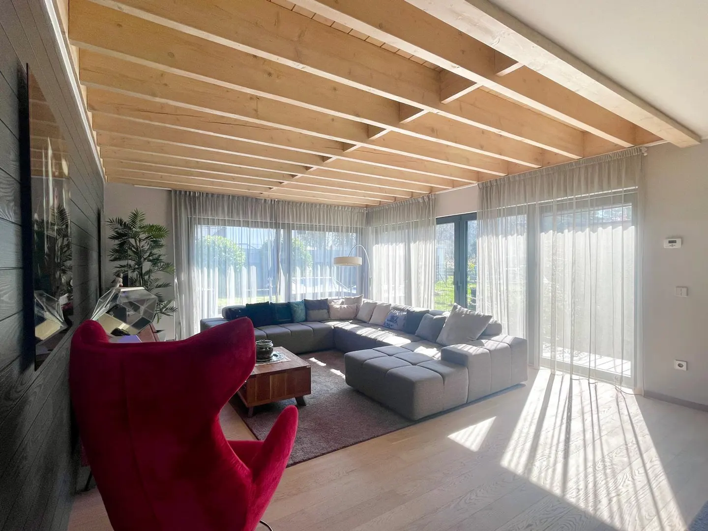 Bright living room with exposed wood beams, large gray sectional sofa, and red accent chair. Sunlight streams through sheer curtains.