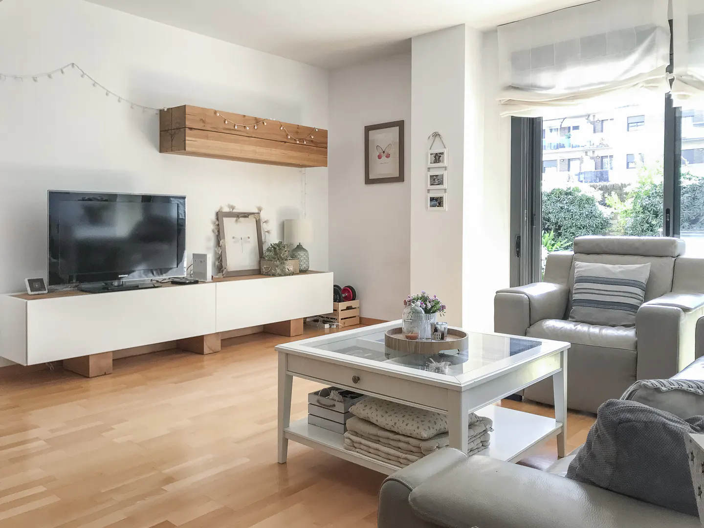 Bright living room with wood floors, white walls, and large windows. A white media console sits below a TV and a wooden shelf. A white coffee table is in front of a gray armchair.