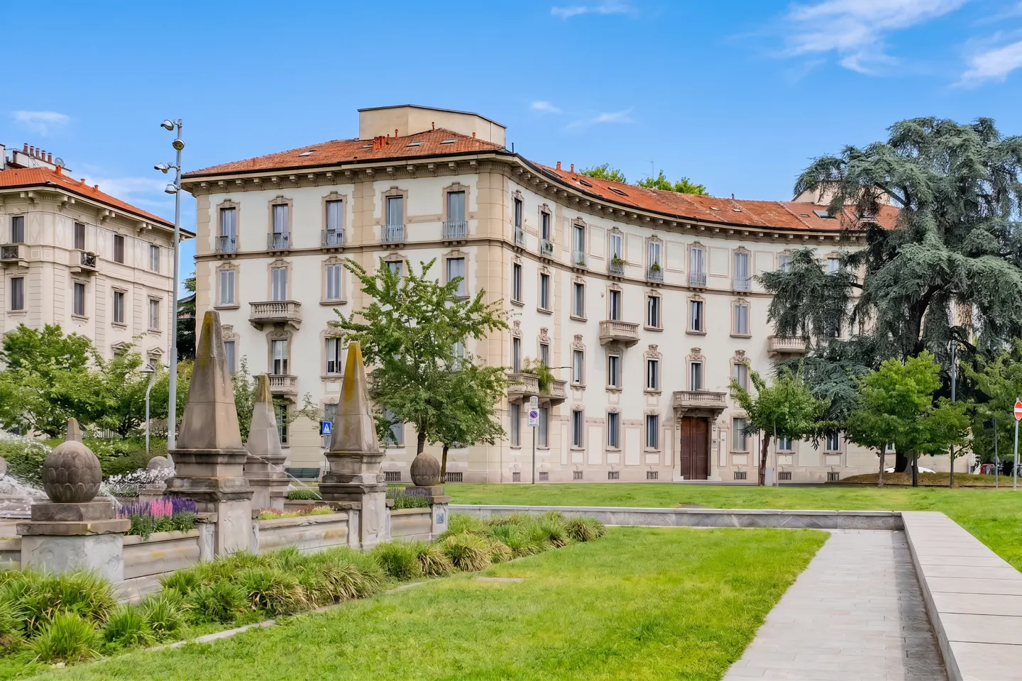 A cream-colored building with a red tile roof overlooks a green lawn and stone planters under a blue sky.