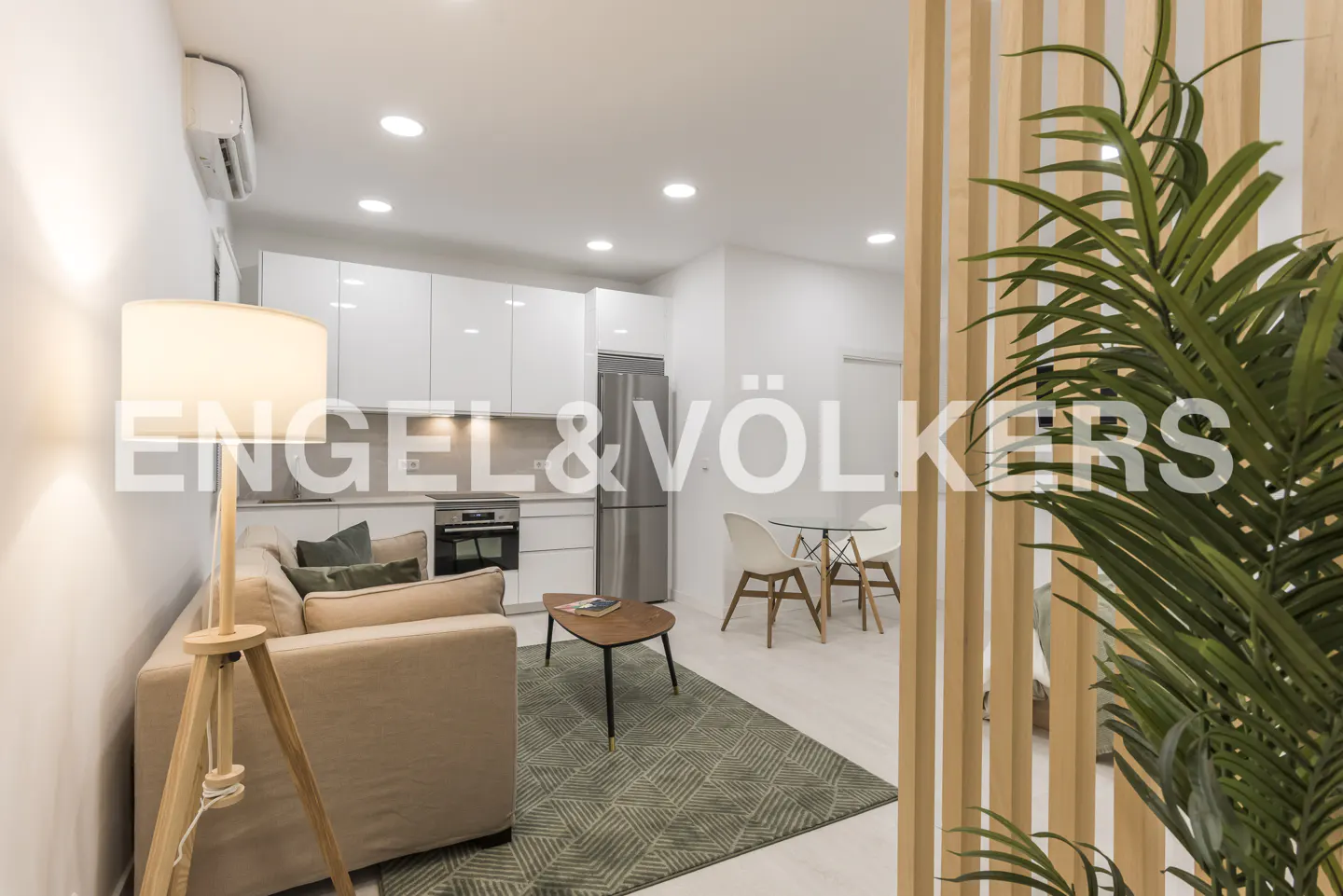 Bright, modern apartment with a beige sofa, green rug, and white kitchen. A wooden screen and plant add texture.