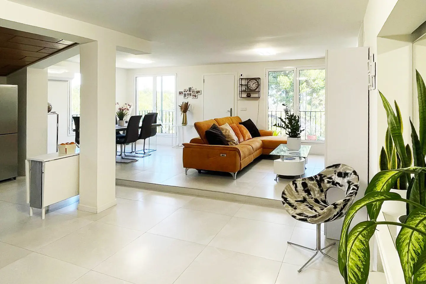 Open-concept living space with white tile floors, a brown sectional sofa, and a dining area with black chairs. Large windows offer natural light.