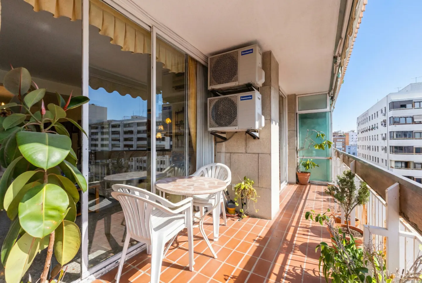 Balcony with red tile floor, white table and chairs, plants, and city view. Two AC units are mounted on the wall.