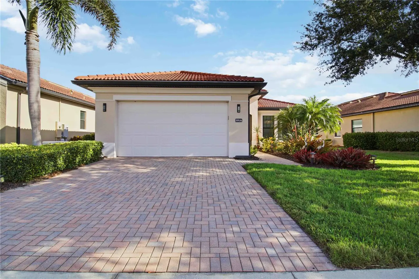 Single-story beige home with a white garage door, red tile roof, brick paver driveway, and lush tropical landscaping.