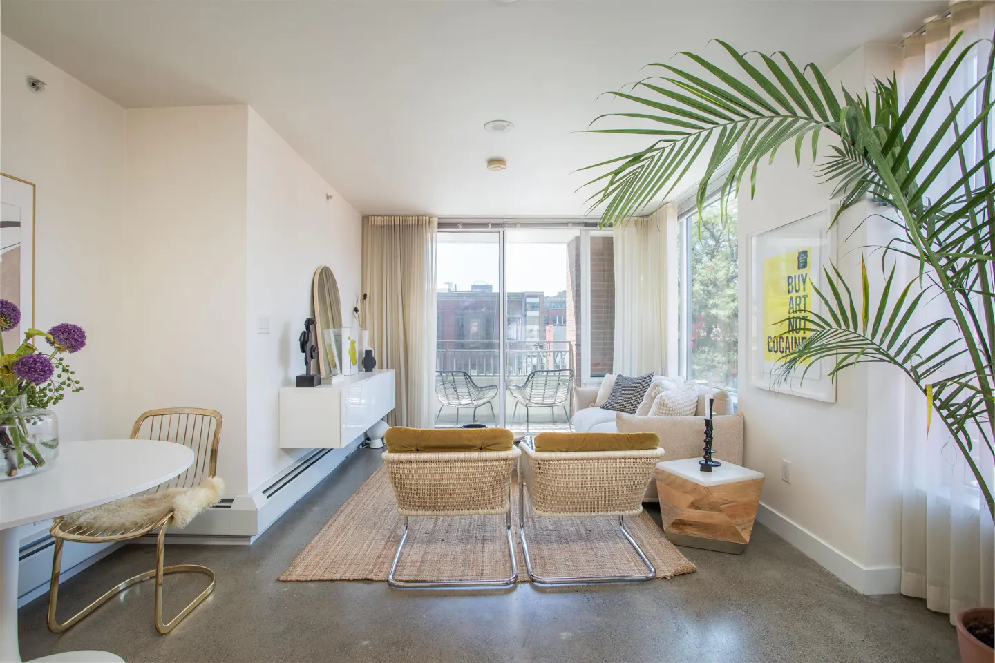 Bright living room with white walls, concrete floors, and a jute rug. Wicker chairs face a balcony with sheer curtains. A palm tree adds a touch of nature.