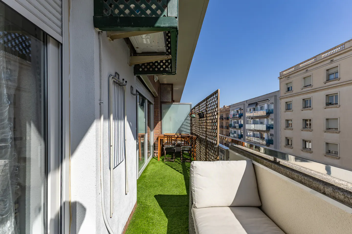 A sunny balcony with artificial grass, a white cushioned bench, and a wooden table with chairs. Buildings are visible in the background.