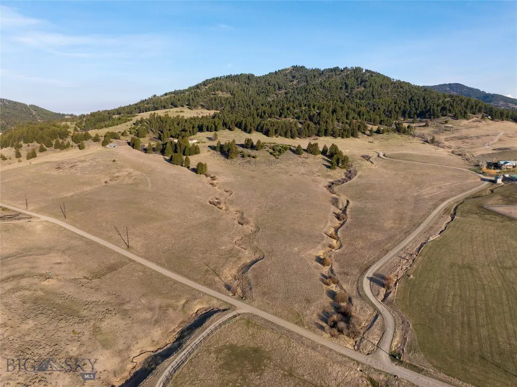 Aerial view of a Montana landscape with brown fields, a winding dirt road, and a forested hill under a blue sky.