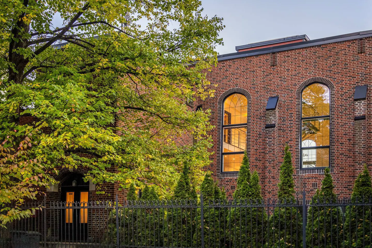 Brick building with arched windows and a black metal fence. Green trees are in the foreground.