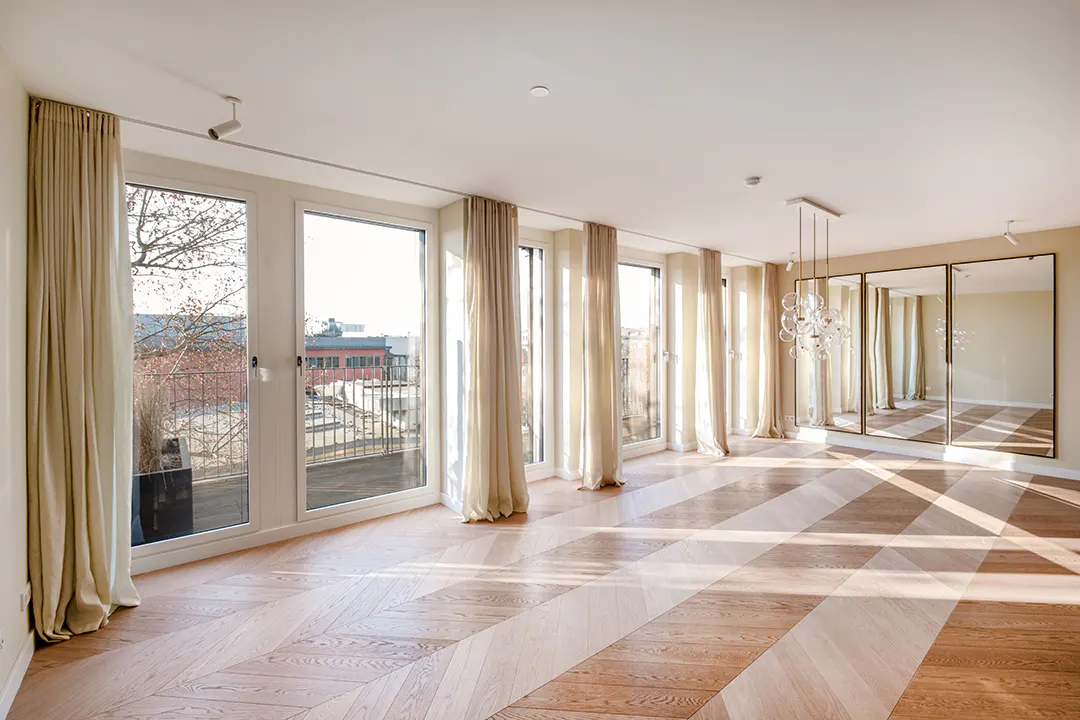 Bright, empty room with herringbone wood floors, beige curtains, and three large mirrors. Windows overlook a balcony and city buildings.