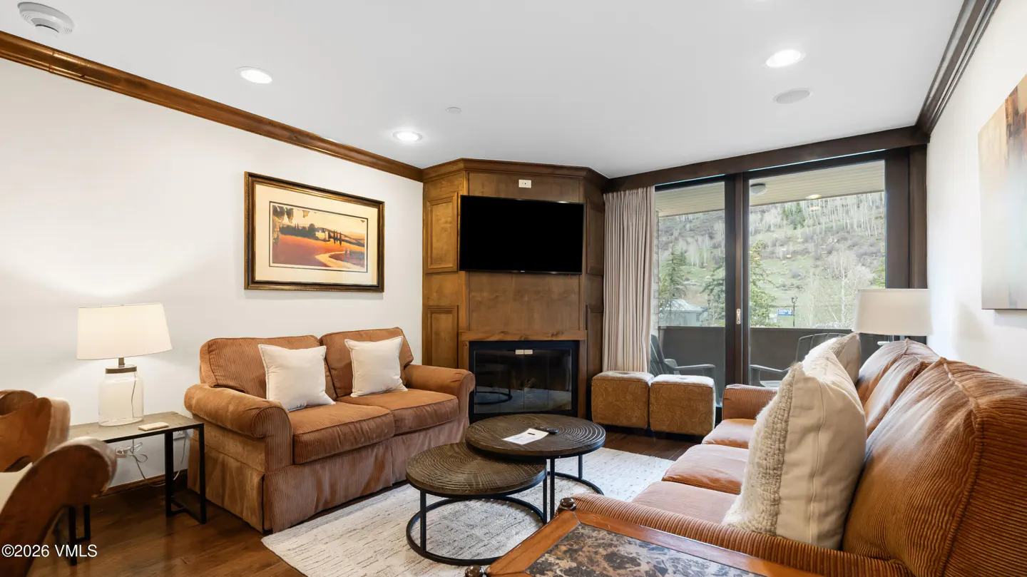 Living room with brown sofas, fireplace, TV, and balcony with mountain view. Two round tables sit on a white rug.