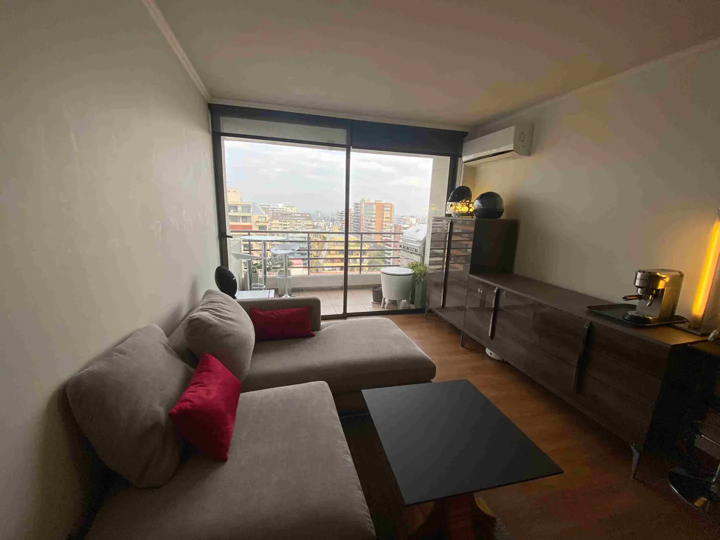 Living room with a gray sectional sofa, red pillows, black coffee table, and a balcony with a city view.