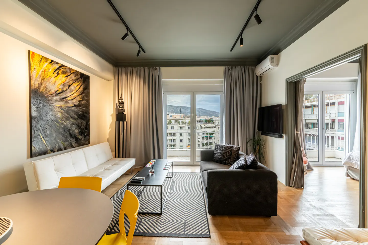 Bright living room with white sofa, gray sofa, yellow chairs, and a large painting. Balcony doors open to a city view.