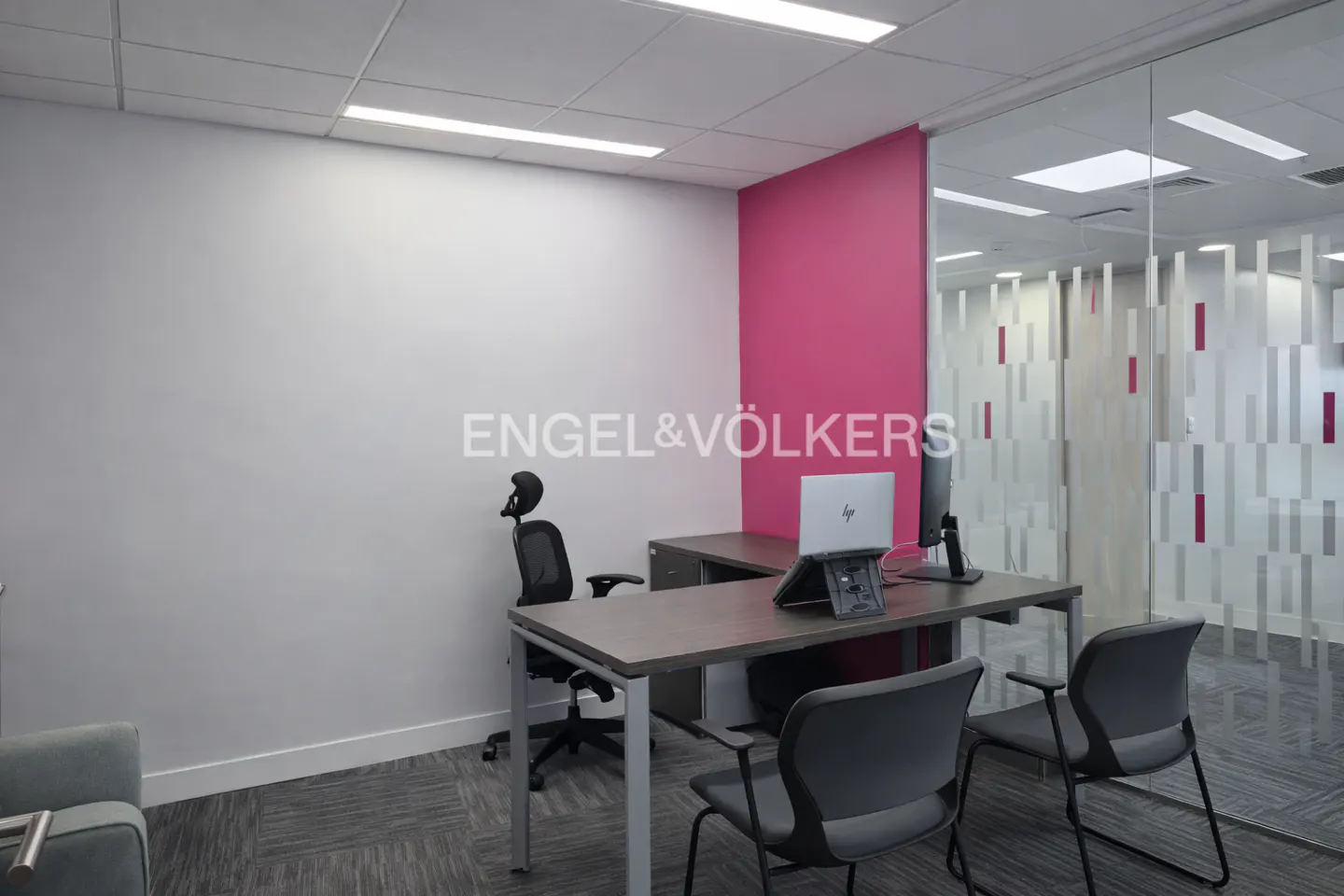 Office space with desk, chairs, and laptop. A pink wall and glass partition add color and light to the room.