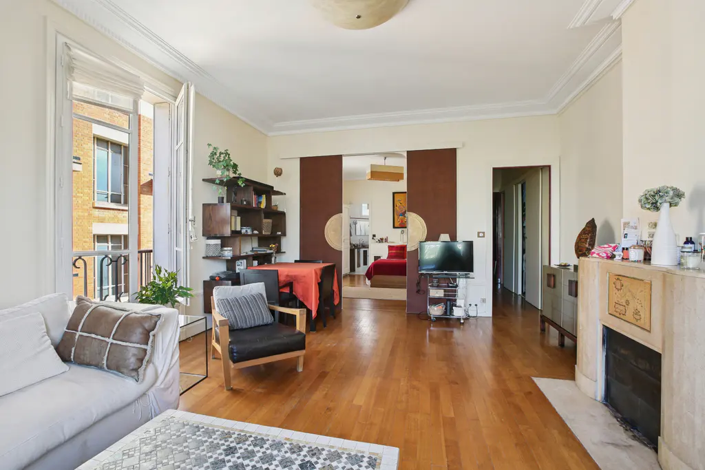 Bright living room with hardwood floors, white sofa, black chair, and open balcony. Brown sliding doors lead to a bedroom.