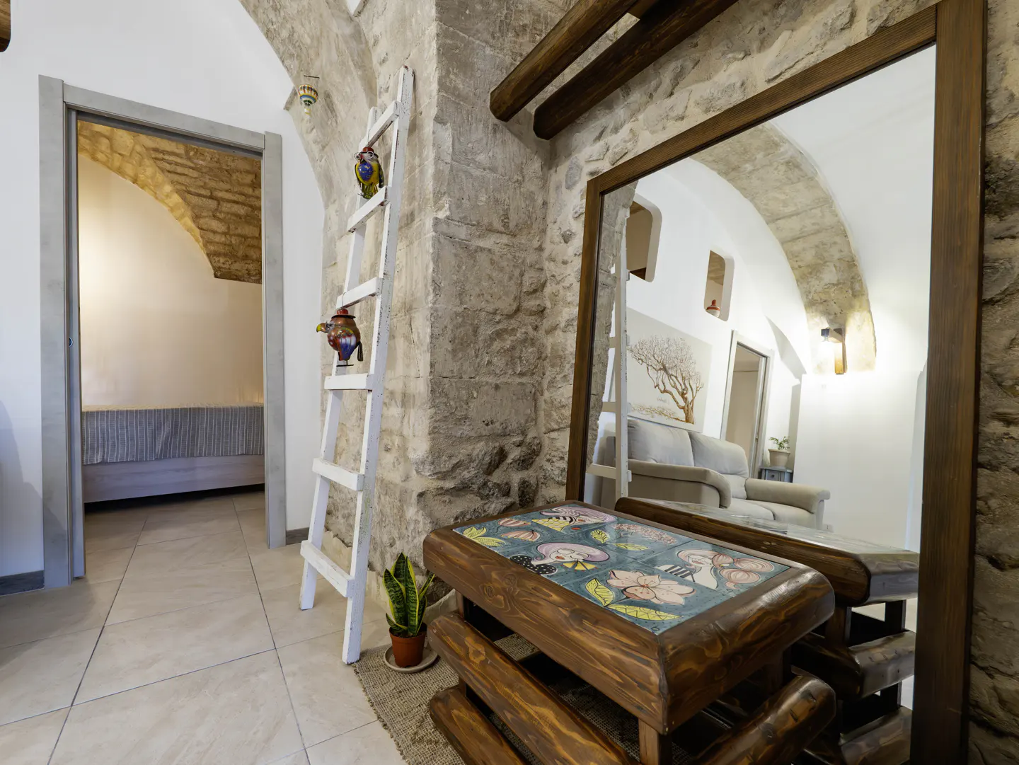 Interior view of a stone-walled room with a mirror reflecting a living area, a white ladder with decorative objects, and a wooden table with a tiled top.