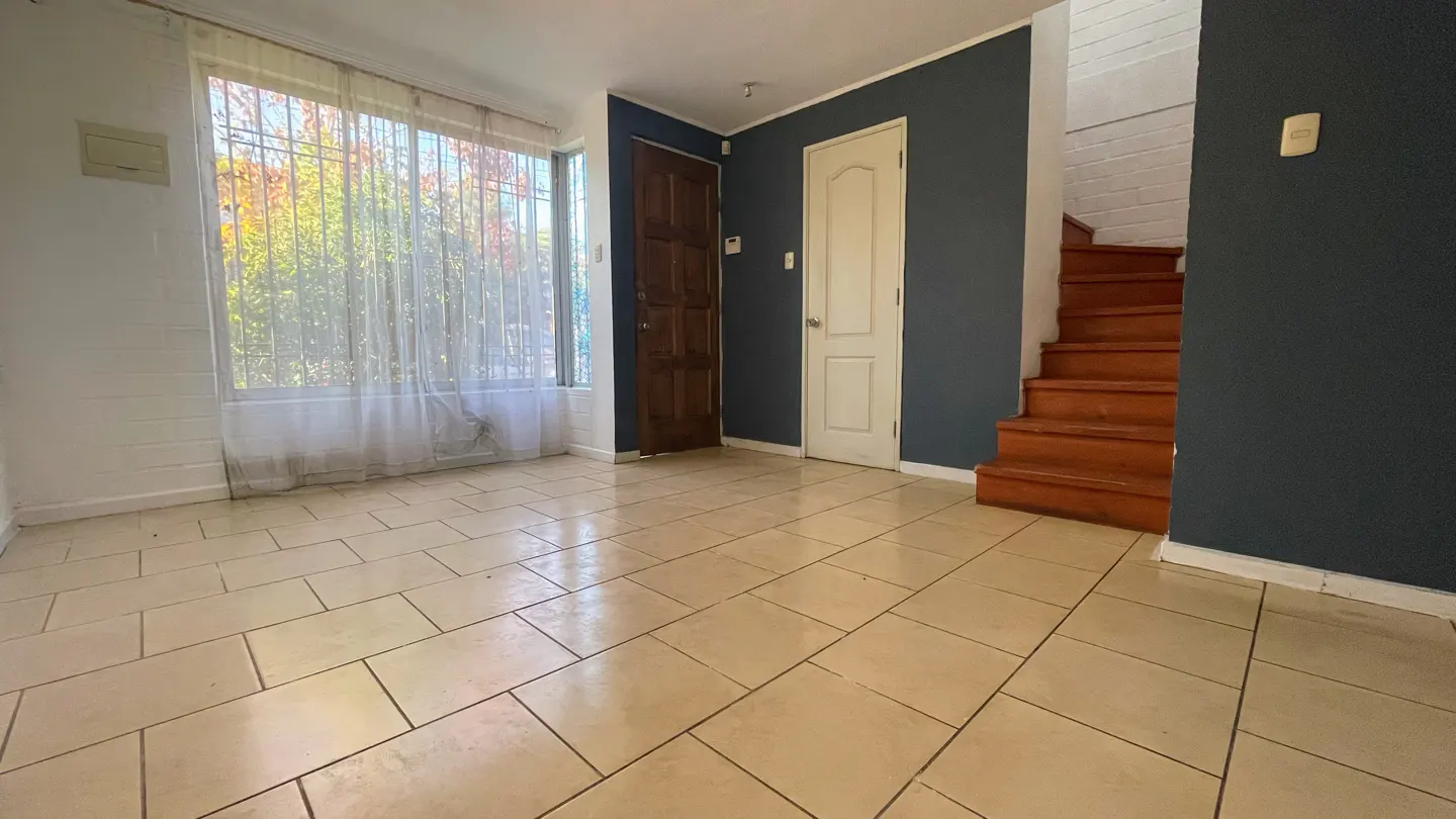 A bright living room with beige tile floors, a large window with sheer curtains, and a wooden staircase.