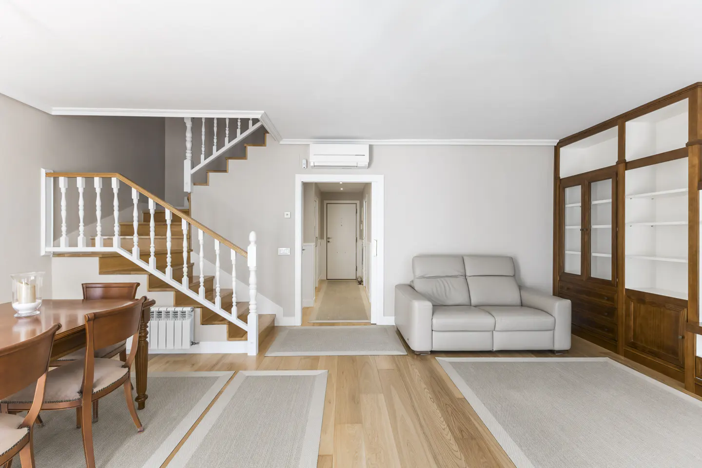 Bright living room with wood floors, a gray sofa, and a wooden dining table. A staircase with white railings leads to the upper floor.