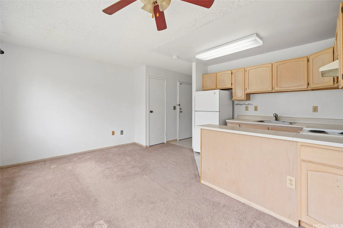 A bright apartment interior with beige carpet, white walls, and light wood cabinets. A red ceiling fan is visible.