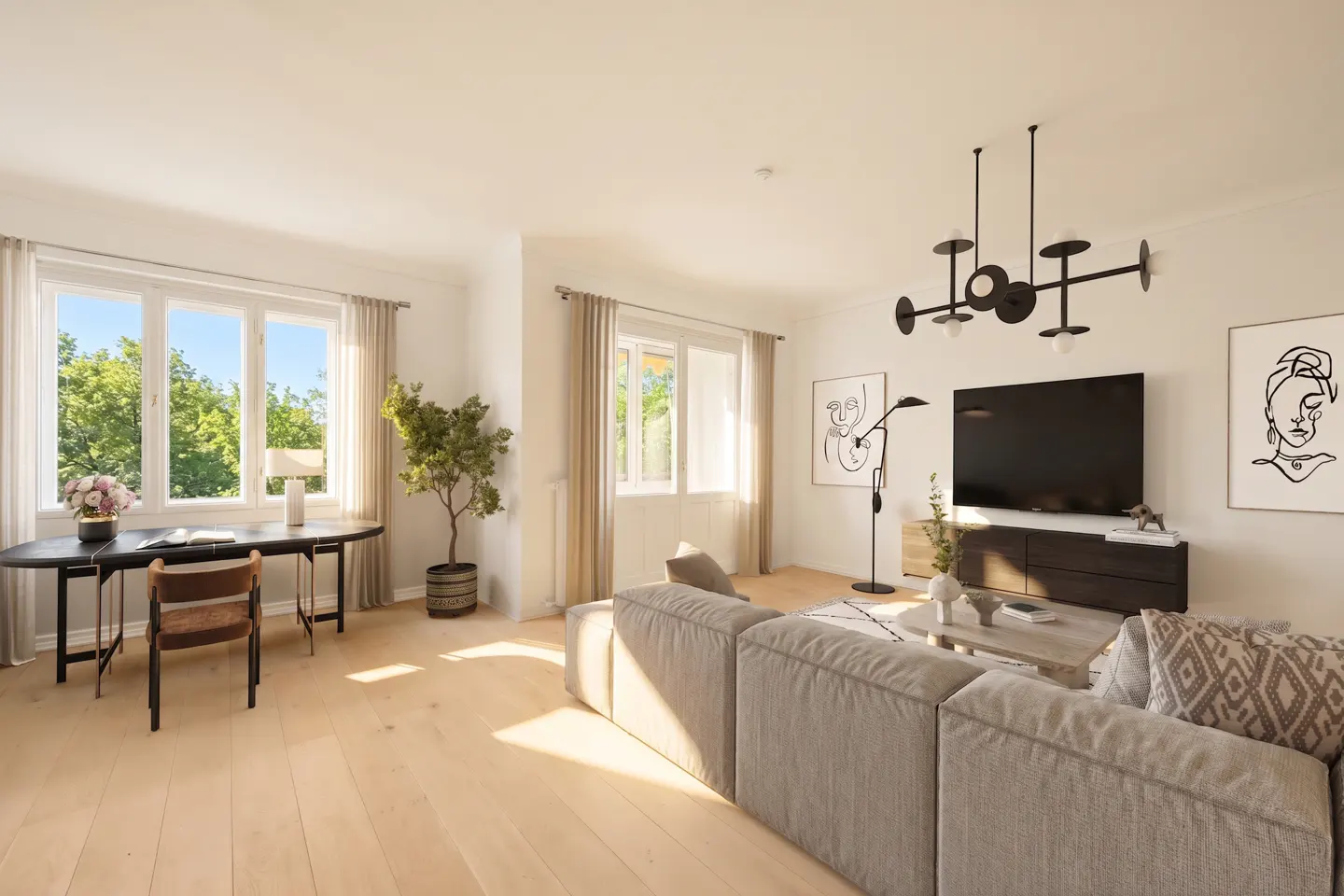 Bright, modern living room with light wood floors, a gray sofa, and a black desk by a window. A black chandelier hangs above a TV.