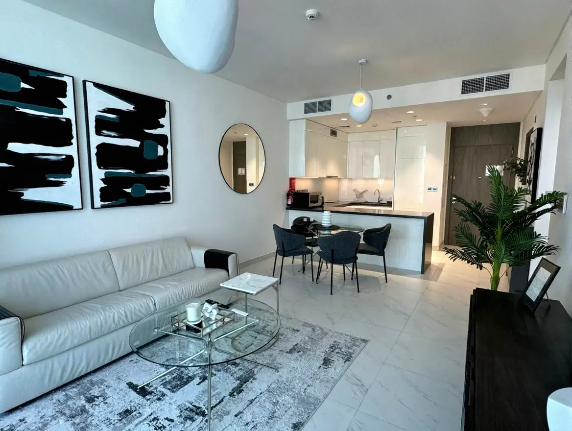 Bright, modern apartment interior with a white sofa, glass table, and open kitchen. Abstract art and a round mirror decorate the walls.