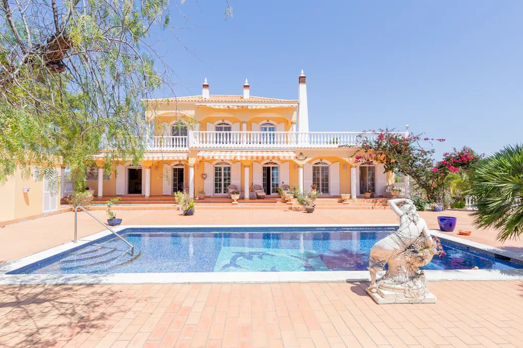 A two-story yellow villa with a swimming pool, a stone statue, and a terracotta patio under a clear blue sky.